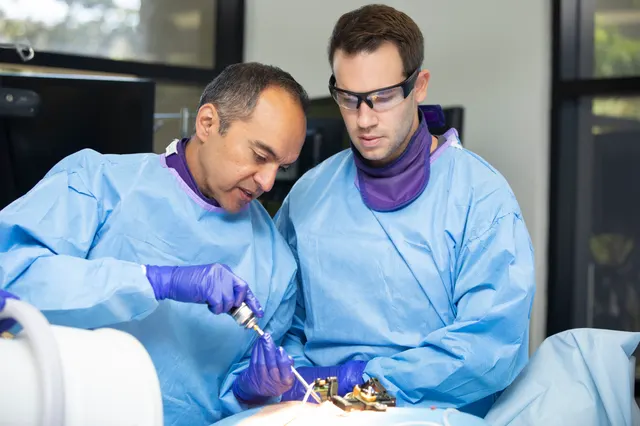An instructor guides a handheld driver at a training station while a course attendee in protective eyewear watches.