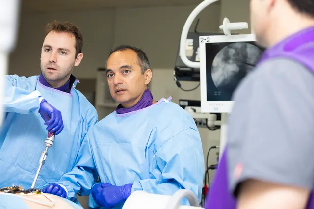 An instructor speaks across a training station to attendees, a fluoroscopy monitor showing a spinal image behind him.