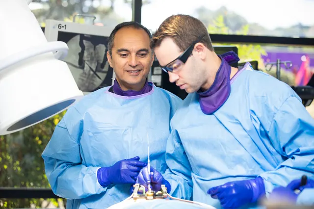 An instructor and an attendee look down at screw hardware seated in a draped training model beneath a surgical light.
