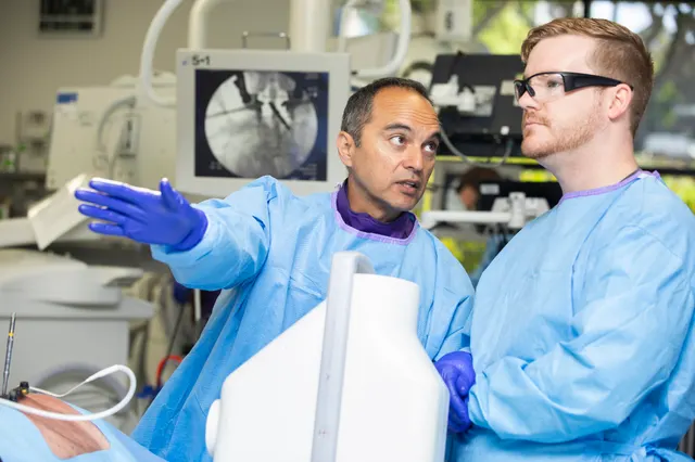 The instructor gestures toward a fluoroscopy monitor showing a screw trajectory while an attendee follows the explanation.