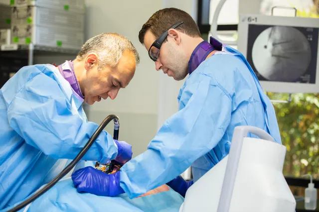 An instructor and an attendee lean over a training station with a powered driver in use, a fluoroscopy image on the monitor behind them.