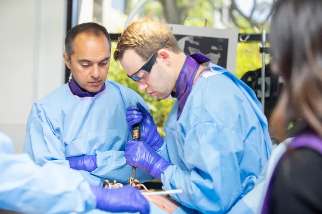 Two clinicians drive hardware into a draped training station while an attendee watches from the foreground.