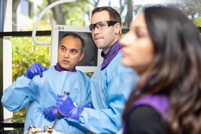 The instructor gestures while explaining a step, with an attendee and a fluoroscopy monitor behind him and another attendee in the foreground.