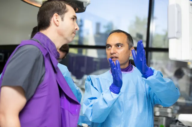 The instructor, gloved hands raised, explains a technique to an attendee wearing a protective lead apron.
