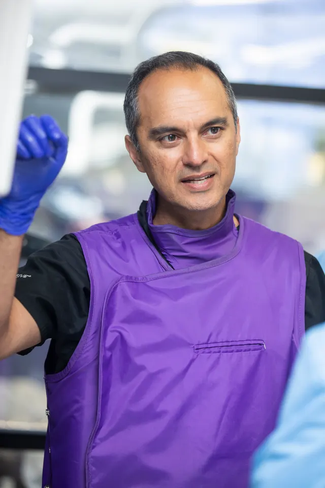 The instructor, wearing a lead apron, speaks to attendees with one gloved hand raised.