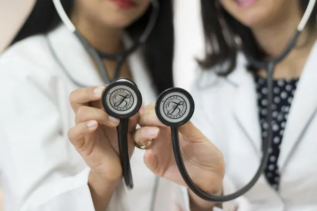 Two clinicians in white coats holding stethoscopes out towards the camera