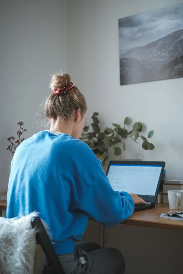 A person seated at a desk, seen from behind, leaning in over a laptop with their shoulders rounded forward
