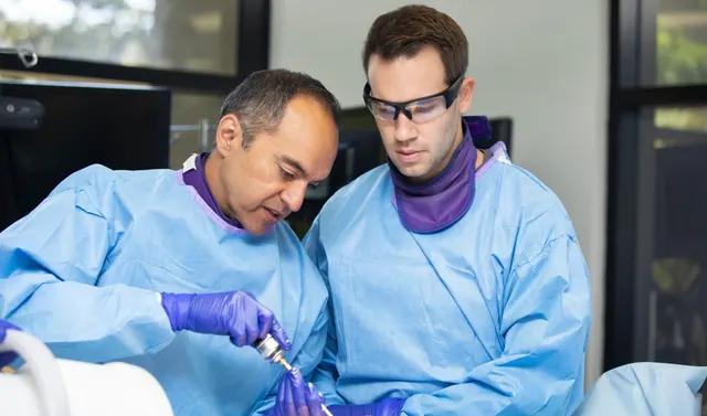 Two clinicians in surgical gowns and gloves at a hands-on training station, one guiding a powered instrument while the other watches
