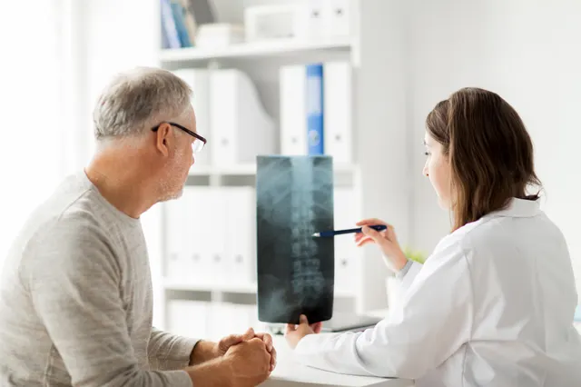 A clinician points to a lumbar spine X-ray while discussing it with an older patient in a clinic room