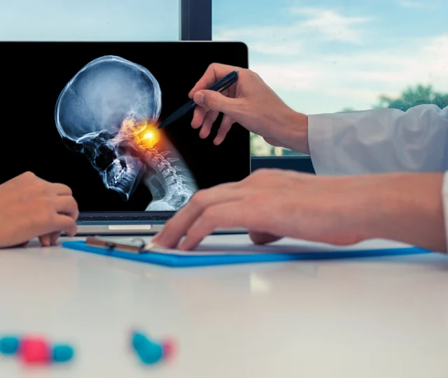 A clinician points to a cervical spine X-ray on a laptop screen, the neck highlighted, while talking with a patient across the desk