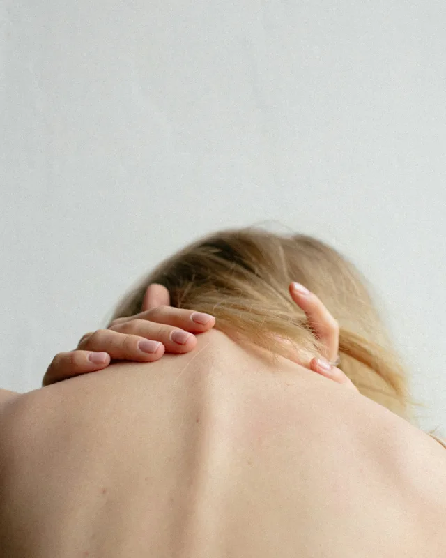 A woman seen from behind, both hands pressed against the back of her neck