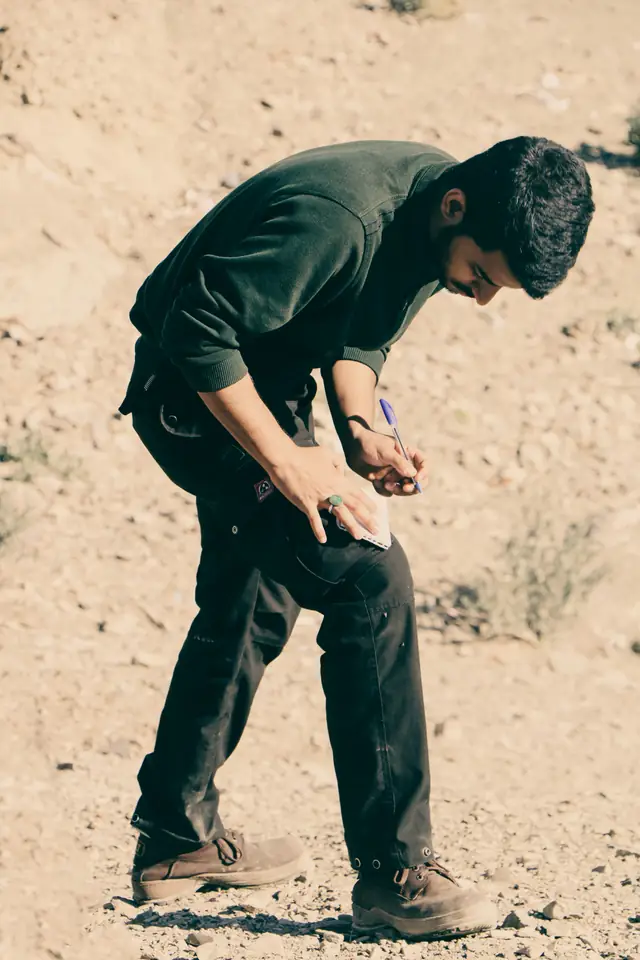A man in work clothes and boots bent sharply forward at the waist on rough stony ground, writing on a small notepad