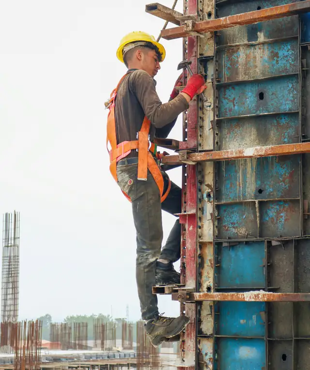 A worker in a hard hat, safety harness and gloves climbing the outside of steel column formwork on a building site