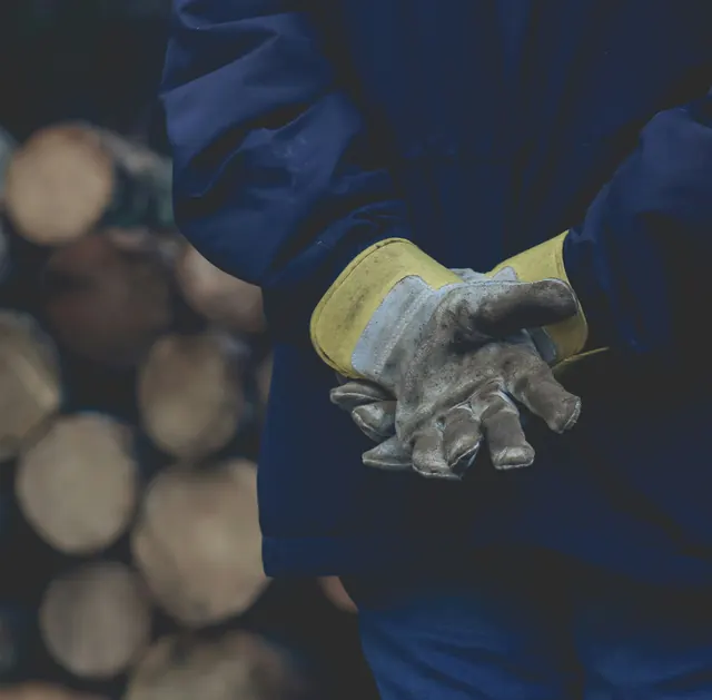A worker seen from behind in heavy leather gloves, hands clasped at the small of the back, beside a stack of cut logs