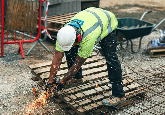 A construction worker in a hi-vis vest and hard hat, bent double at the waist, cutting steel reinforcing mesh with an angle grinder