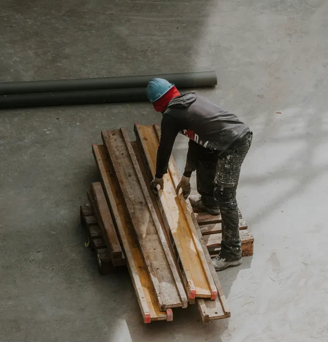 A worker in a hard hat, seen from above, bending forward over a stack of long timber planks on a bare concrete floor